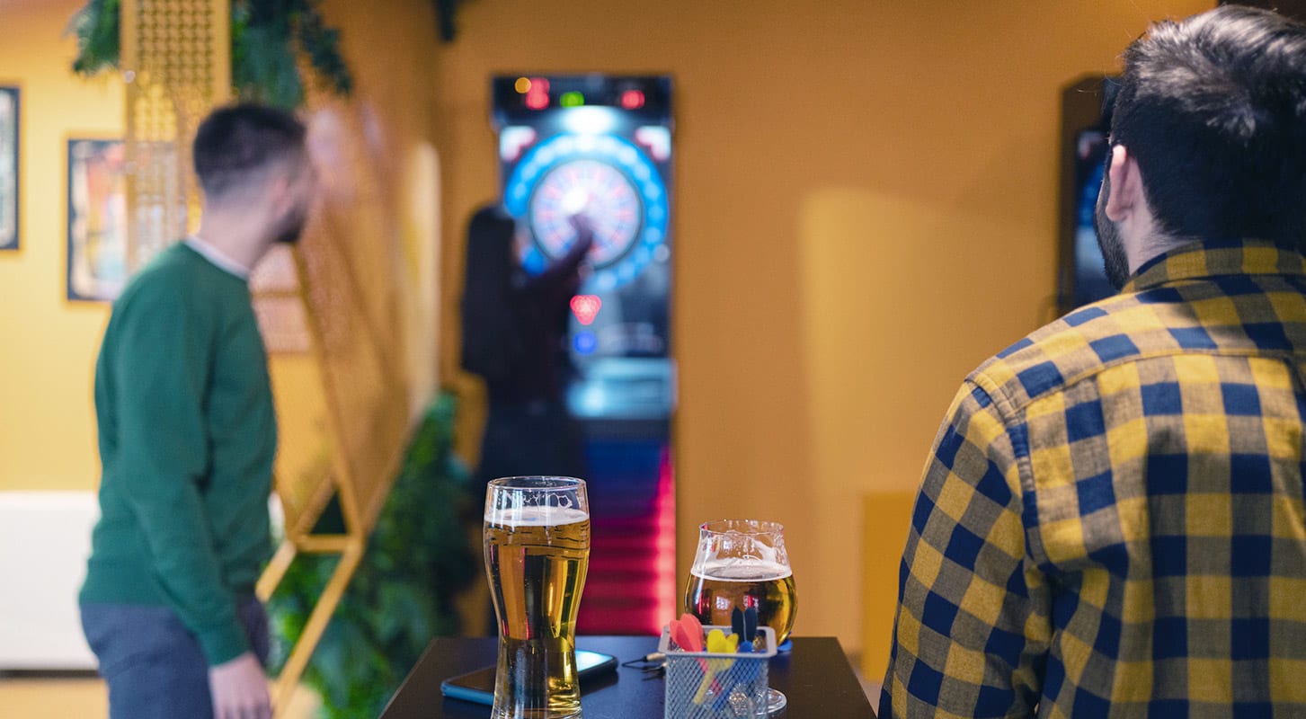 Two men engaged in a game of darts at a bar, with drinks and a dartboard in the background.